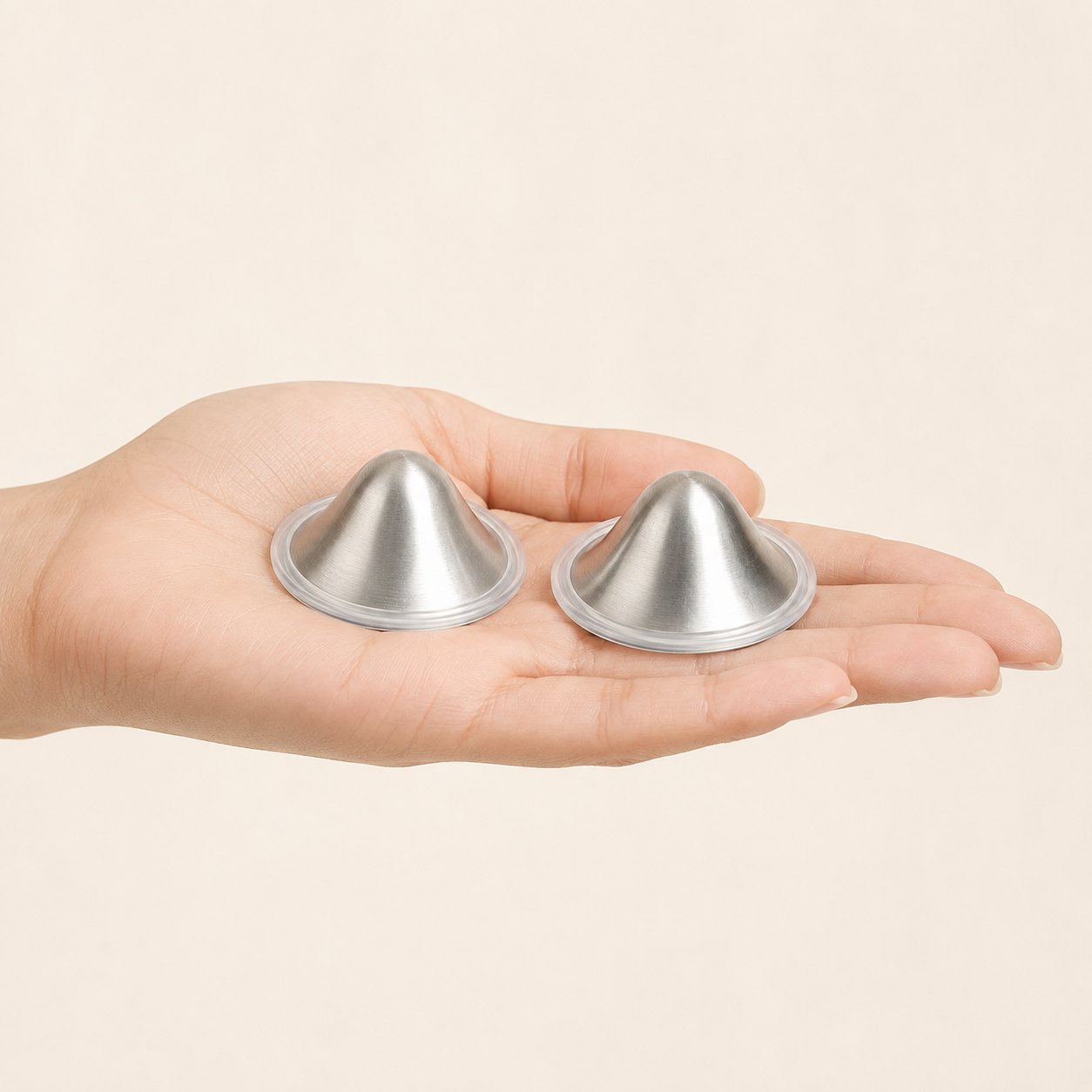 A pair of small silver nursing cup domes resting across a mother's open fingers, showing their true scale
