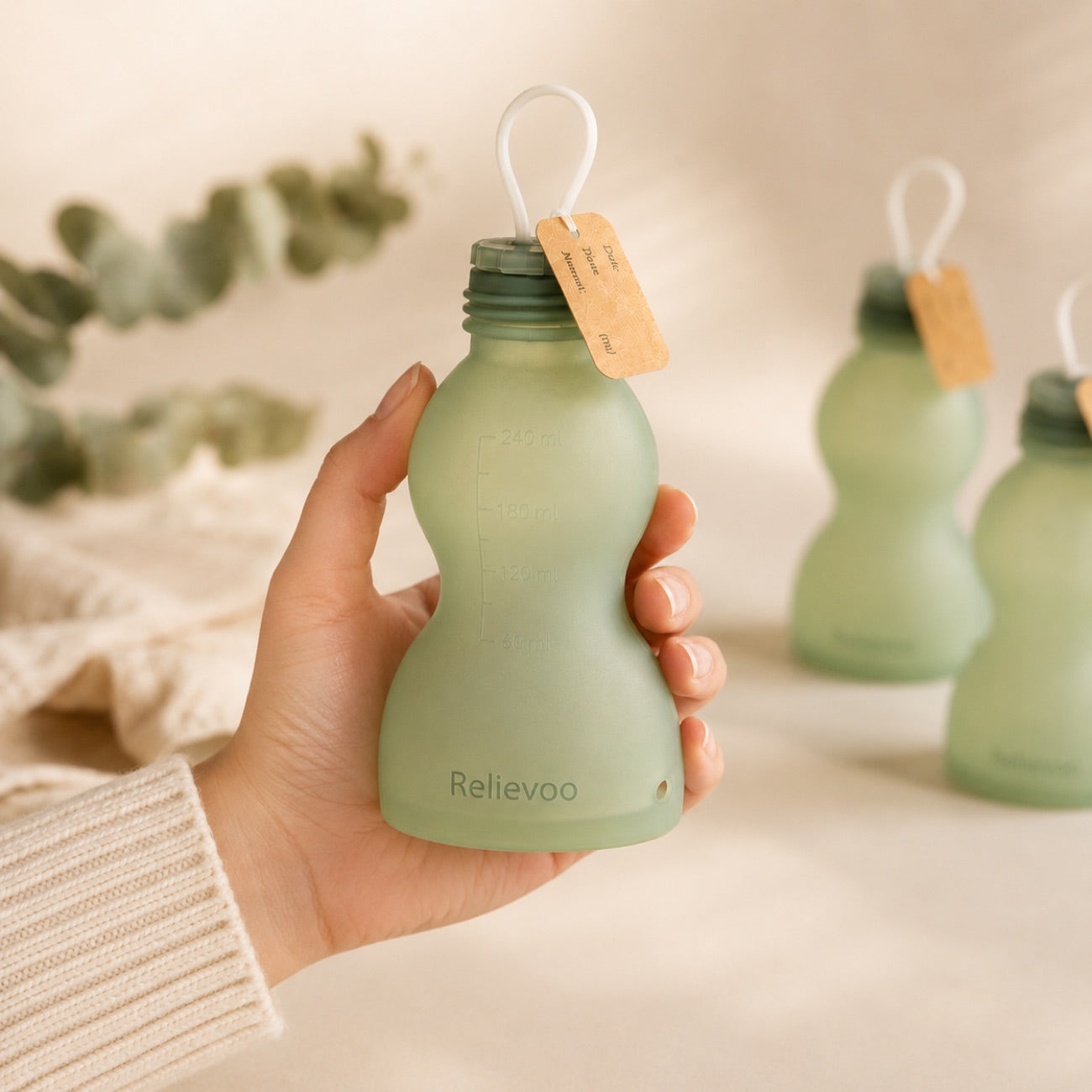 Hand holding a Relievoo reusable sage-green silicone breast milk storage bottle showing its size and measurement markings, with more bottles behind